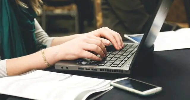 Hands typing on a laptop, with a notebook and phone on the desk