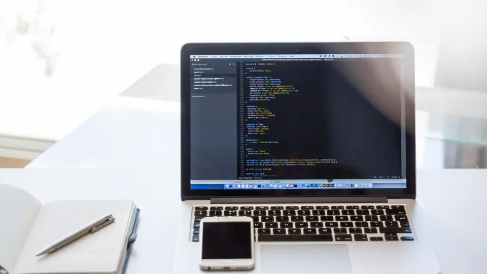 Laptop showing code, with a phone and notebook on the desk